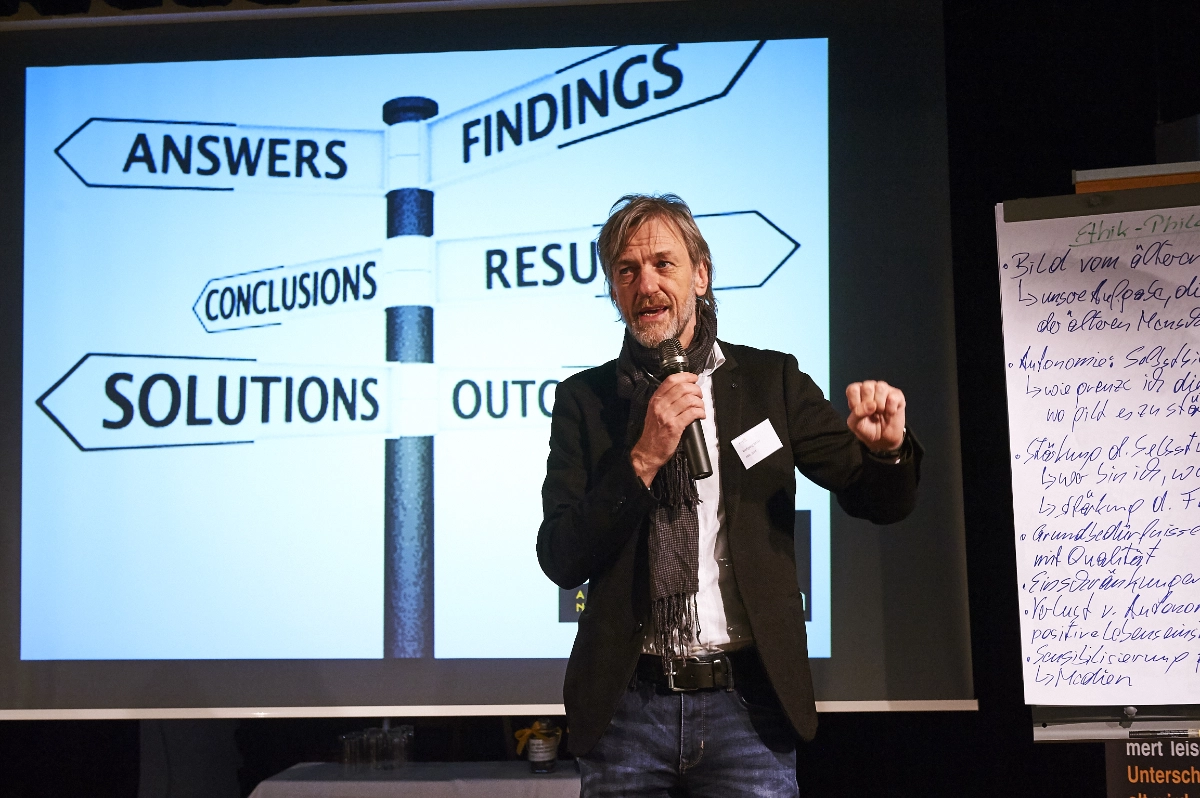 Wolfgang Rath spricht bei einem Fachvortrag mit Mikrofon vor einer Präsentationsfolie und einem Flipchart.