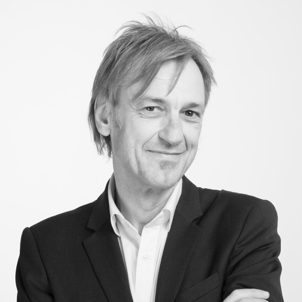Schwarzweiß-Porträt von Wolfgang Rath mit hellem Hemd und dunklem Sakko vor hellem Hintergrund, die Arme verschränkt.