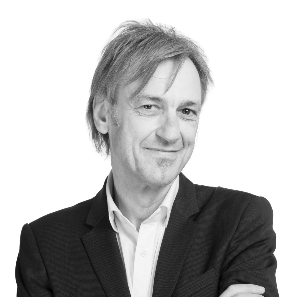 Schwarzweiß-Porträt von Wolfgang Rath mit hellem Hemd und dunklem Sakko vor hellem Hintergrund, die Arme verschränkt.