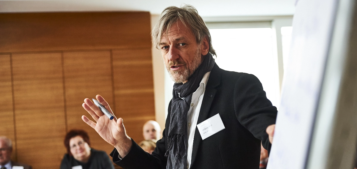 Wolfgang Rath steht vor einem Flipchart, spricht zu einer Gruppe von Teilnehmenden und hält einen Stift in der Hand.