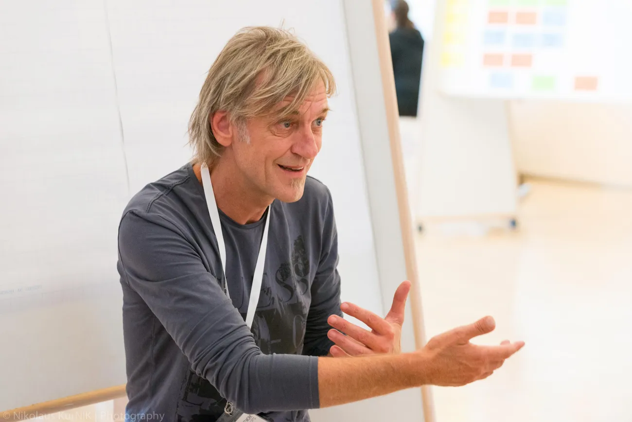 Wolfgang moderiert einen Workshop und erklärt mit lebhaften Gesten einen Sachverhalt vor einem Flipchart.